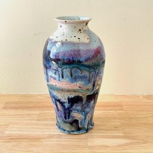 May include: A ceramic vase with a narrow neck and a flared rim, featuring a glossy glaze with a drip pattern in shades of deep blue, purple, teal, and soft pink. The upper portion is off-white with brown speckles, transitioning into colors.