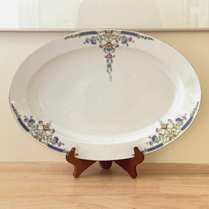 May include: An oval white ceramic serving platter featuring a decorative border of blue, pink, and green floral patterns with gold accents. The design includes symmetrical botanical motifs along the rim and a central floral drop at the top edge.