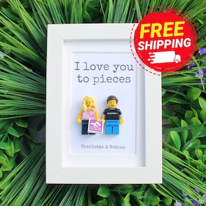 May include: A white rectangular frame containing a white background with the printed text I love you to pieces and Charlotte & Nathan. Two plastic toy figures stand in the center, one with blonde hair and one with brown hair, holding a small sign.
