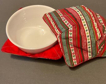 May include: A white ceramic bowl rests on a red quilted bowl cozy. A second bowl cozy with a red, green, and white striped pattern is next to the bowl. The cozy has a festive design with candy cane and snowflake motifs.