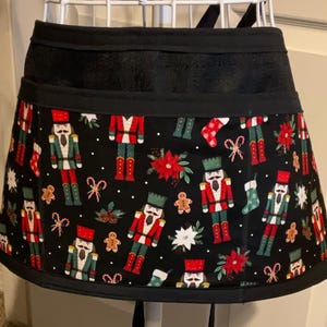 May include: A black Christmas-themed apron with a festive pattern. The apron features a black waistband and ties, and the lower portion is adorned with nutcrackers, gingerbread men, poinsettias, and candy canes. The apron is designed for kitchen use.