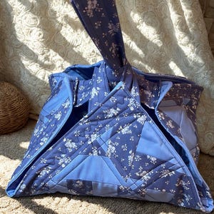 May include: A quilted tote bag in shades of blue, featuring a floral pattern. The bag has a unique geometric design with a handle. The fabric appears to be cotton, and the bag is sitting on a textured surface.