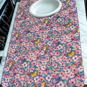 May include: A rectangular fabric bib featuring a blue background with a repeating pattern of pink cherry blossoms and yellow butterflies. The bib has a white, oval-shaped neck opening positioned near the top center of the fabric panel.