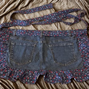 May include: A denim apron with a floral patterned trim and straps. The apron features a dark blue denim base with two back pockets. The trim and straps are navy blue with a repeating pattern of red, white, and blue flowers.