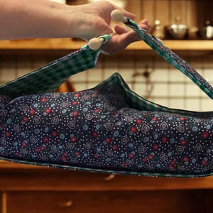 May include: A fabric carrier featuring a dark blue exterior with a white polka dot, red flower, and heart pattern. The interior is lined with a green and white checkered fabric. The carrier includes two wooden beads attached to the top handles.