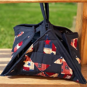May include: A navy blue quilted tote bag with a patriotic patchwork design. The bag features red, white, and blue star and stripe patterns. It has a handle for carrying and is designed to hold items.