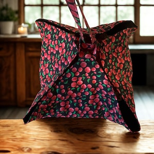May include: A fabric bag featuring a repeating pattern of red cherries and green leaves on a black background. The bag has a square-shaped base with four triangular flaps that fold upward to meet at a central handle, creating a structured shape.