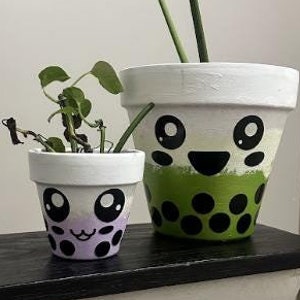 May include: Two white ceramic plant pots with hand-painted cartoon faces. The larger pot has green and black polka dots and a green mouth. The smaller pot has purple and black polka dots and a pink mouth. Both pots have black eyes.