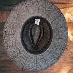 May include: A gray felt fedora hat with a spiderweb design. The hat has a wide brim and a black band around the crown.