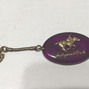 Op de afbeelding: Een vintage sleutelhanger met een goudkleurig paard en jockey op een paarse ovaal. De tekst "Hollywood Park" is in goud. De sleutelhanger heeft een goudkleurige ring en een opgerolde metalen ketting.