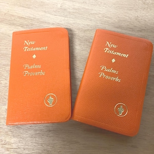 May include: Two bright orange pocket-sized books with textured covers. Each cover features gold embossed text reading New Testament, Psalms, and Proverbs, with a small gold diamond symbol and a circular emblem at the bottom center.