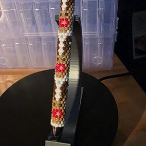 May include: A handcrafted pen with a beaded design. The pen features a pattern of white, brown, and red beads. The pen is displayed on a black stand, showcasing the intricate beadwork and craftsmanship.