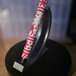 May include: A decorative pen with a white and red beaded design. The pen is displayed on a black stand with a silver tip. The stand is on a black circular base.