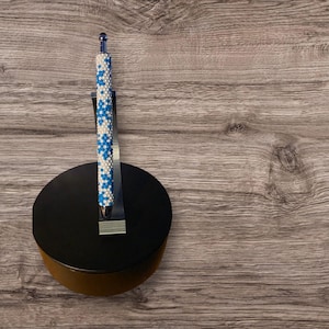 May include: A beaded pen with a white and blue floral pattern. The pen is displayed on a black stand with a silver metal holder. The pen has a clear, rounded top and a silver tip.