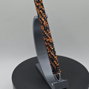 May include: A decorative pen with a silver tip and top, covered in orange and black crystals. The pen is displayed on a gray stand, set against a white background. The pen is a writing instrument.
