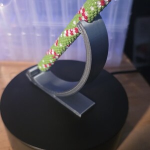 May include: A green, red, and white beaded pen rests on a black stand. The pen has a silver tip and a decorative pattern. The stand is round with a curved support.