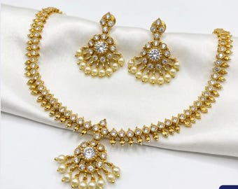 May include: A gold-toned necklace and matching earring set featuring clear round-cut stones and small cream-colored pearls. The necklace has a central pendant with a large stone, while the earrings mirror this fan-shaped design.