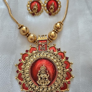 May include: A gold-toned necklace and matching earrings set. The necklace features a large pendant with a red center and a detailed gold figure. The earrings have a similar design. The set is displayed on a white surface.