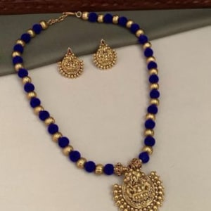 May include: A handcrafted necklace and earring set. The necklace features navy blue pom-poms and gold-colored beads. The pendant and earrings have a detailed, ornate design. The jewelry set is displayed on a neutral surface.