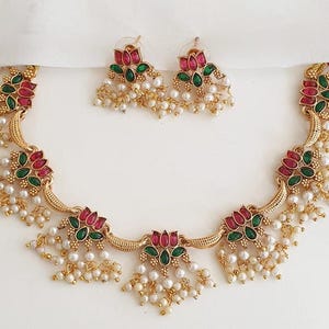 May include: A gold-toned necklace and matching earring set featuring lotus-shaped motifs inlaid with pink and green stones. The jewelry is accented with clusters of small white pearls hanging from the base of each floral element and along the chain.