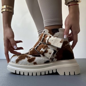 May include: Cow print ankle boots with a white sole and two buckle closures. The boots have a brown and white pattern and a thick, ridged sole. The boots are being adjusted by a person wearing gold bracelets and a watch.