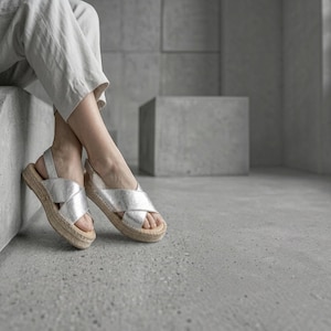 May include: A pair of metallic silver sandals featuring wide crisscross straps over the top of the foot, an adjustable ankle strap with a buckle, and a textured jute-wrapped platform sole. The sandals are worn with light-colored linen trousers.