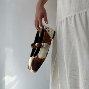 May include: A pair of Mary Jane style shoes with a cowhide pattern in brown and white. The shoes have a strap with a silver buckle and a black sole. The shoes are held by a person.