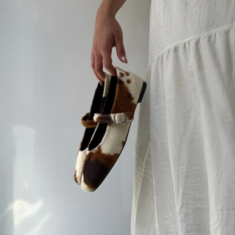 May include: A pair of Mary Jane style shoes with a cowhide pattern in brown and white. The shoes have a strap with a silver buckle and a black sole. The shoes are held by a person.