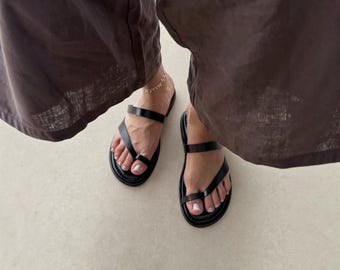 May include: A pair of black leather sandals featuring a toe loop design and a secondary horizontal strap across the midfoot. The sandals have a flat, rounded sole and are worn with dark brown wide-leg trousers and a delicate silver ankle bracelet.
