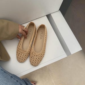 May include: A pair of beige woven ballet flats featuring a lattice pattern, a small decorative bow on the rounded toe, and a smooth inner sole with the brand name MUPA printed in black. The shoes have a low-profile design and a neutral tan color.