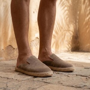 May include: A pair of brown suede espadrille slip-on shoes. The shoes have a rounded toe and a woven jute sole. The image shows the shoes being worn by a person.