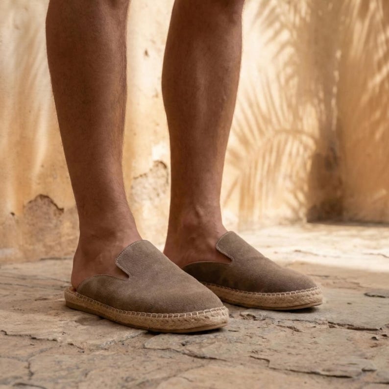 May include: A pair of brown suede espadrille slip-on shoes. The shoes have a rounded toe and a woven jute sole. The image shows the shoes being worn by a person.