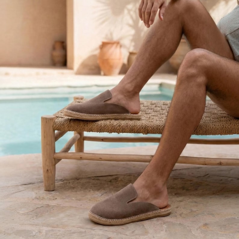 May include: Taupe suede slip-on shoes with a woven jute sole. The open-back design offers a relaxed fit. The shoes are shown on a person's feet, suggesting casual wear. The neutral color makes them versatile for various outfits.