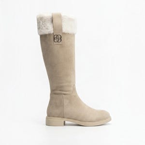 May include: A knee-high boot in a light beige color, featuring a soft white faux fur cuff at the top. A vertical beige strap runs down the side, accented with a metallic silver-toned logo emblem. The boot has a flat sole and a rounded toe design.