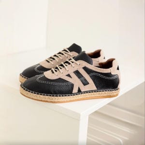 May include: A pair of black and beige sneakers with a textured leather upper and a beige suede accent. The shoes feature white stitching and a woven jute sole. The laces are beige. The sneakers are displayed on a white surface.