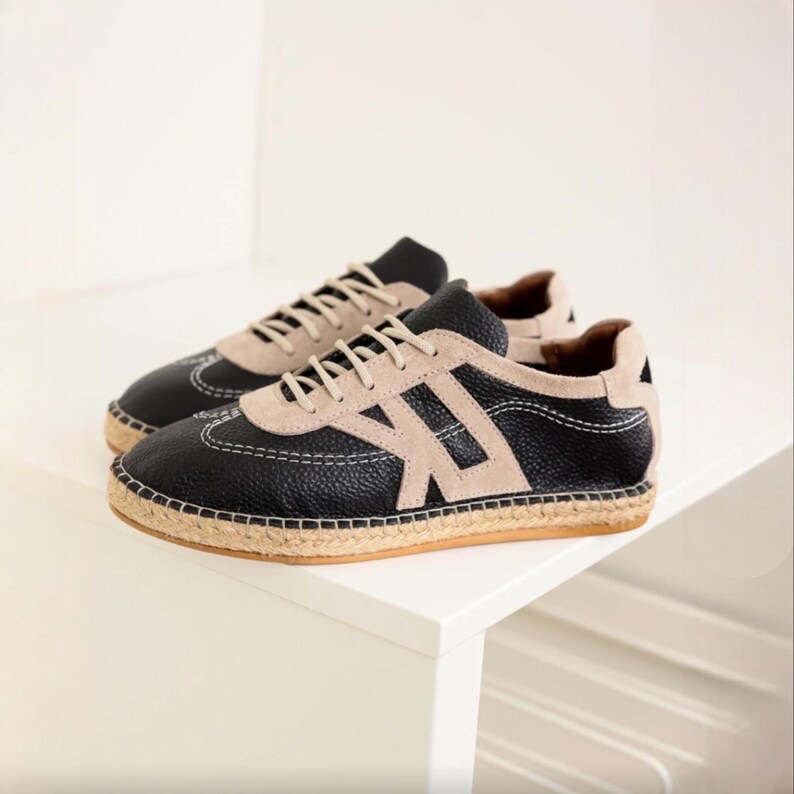 May include: A pair of black and beige sneakers with a textured leather upper and a beige suede accent. The shoes feature white stitching and a woven jute sole. The laces are beige. The sneakers are displayed on a white surface.
