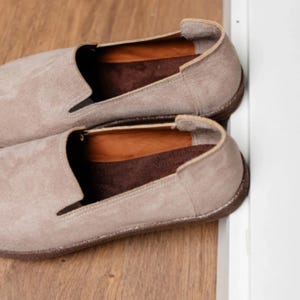 Puede incluir: Un par de zapatos sin cordones de ante beige. Los zapatos tienen una puntera redondeada y una suela marr&oacute;n oscuro. El interior est&aacute; forrado con un material marr&oacute;n m&aacute;s oscuro. Los zapatos est&aacute;n colocados sobre una superficie de madera.