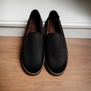 Puede incluir: Un par de zapatos negros sin cordones con punta redonda y suela marr&oacute;n. Los zapatos est&aacute;n hechos de un material mate y se exhiben sobre una superficie de madera. El interior del zapato es marr&oacute;n.