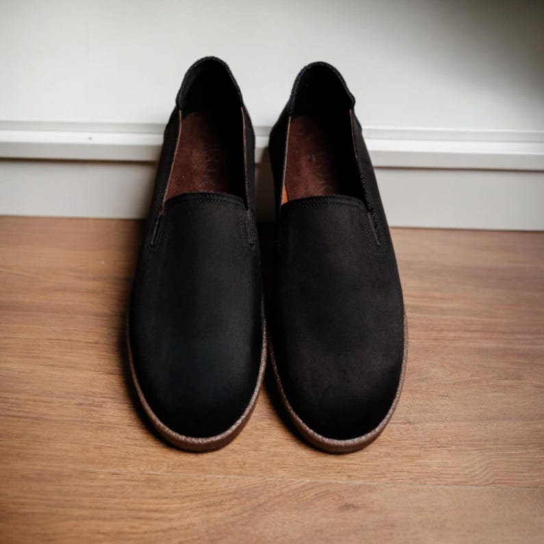 Puede incluir: Un par de zapatos negros sin cordones con punta redonda y suela marr&oacute;n. Los zapatos est&aacute;n hechos de un material mate y se exhiben sobre una superficie de madera. El interior del zapato es marr&oacute;n.