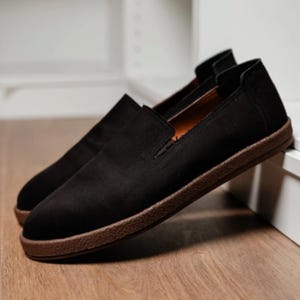 Puede incluir: Un par de zapatos negros sin cordones con suela marr&oacute;n. Los zapatos est&aacute;n hechos de un material suave y tienen un dise&ntilde;o simple y cl&aacute;sico. Los zapatos se exhiben sobre una superficie de madera.