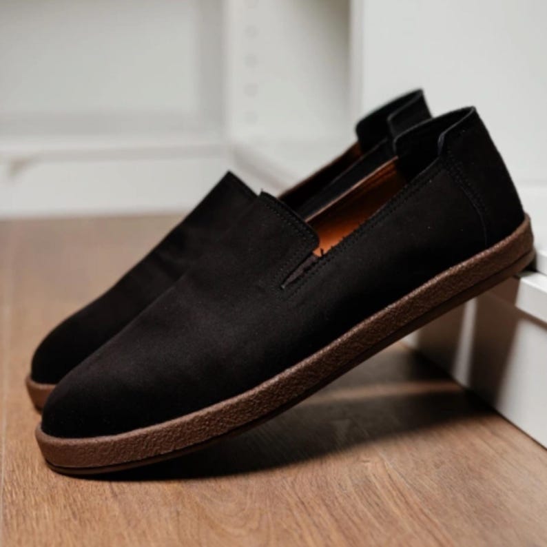Puede incluir: Un par de zapatos negros sin cordones con suela marr&oacute;n. Los zapatos est&aacute;n hechos de un material suave y tienen un dise&ntilde;o simple y cl&aacute;sico. Los zapatos se exhiben sobre una superficie de madera.