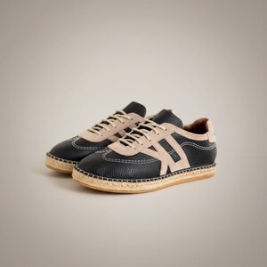 May include: Black leather sneakers with beige suede detailing and a woven jute sole. The trainers have white stitching and beige laces. The design includes a beige letter "A" on the side.