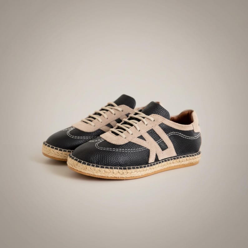 May include: Black leather sneakers with beige suede detailing and a woven jute sole. The trainers have white stitching and beige laces. The design includes a beige letter "A" on the side.