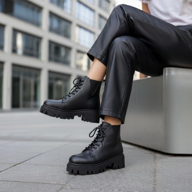 K&ouml;nnte beinhalten: Schwarze Schn&uuml;rstiefel mit klobiger Profilsohle. Die Stiefel haben eine runde Spitze und eine glatte, leder&auml;hnliche Oberfl&auml;che. Die Stiefel werden mit einer schwarzen Hose getragen.