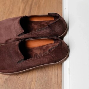 Puede incluir: Un par de zapatos sin cordones de ante marr&oacute;n. Los zapatos tienen una punta redondeada y un perfil bajo. El interior presenta una plantilla de cuero marr&oacute;n. Los zapatos est&aacute;n sobre una superficie de madera.