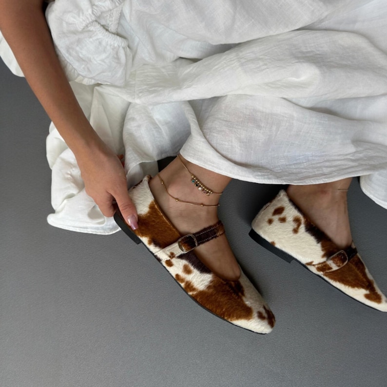 May include: Cowhide print Mary Jane flats with a brown leather strap and buckle. The shoes have a white and brown spotted pattern and a black sole. The shoes are a stylish and unique footwear option.