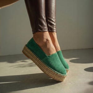 May include: Green suede espadrille platform shoes with a woven jute sole. The shoes have a rounded toe and a slip-on design. The platform sole is approximately 3.8 cm high. The shoes are shown against a neutral background.