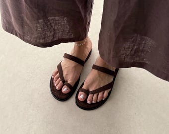 May include: A pair of dark brown flat sandals featuring a toe-loop design and a single horizontal strap across the midfoot. The sandals have a rounded sole with visible stitching along the edge, providing a simple and functional footwear option.