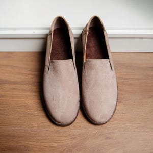 Puede incluir: Un par de zapatos sin cordones de ante beige. Los zapatos tienen una puntera redondeada y un perfil bajo. El interior es de color marr&oacute;n oscuro. Los zapatos est&aacute;n colocados sobre una superficie de madera.