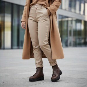 K&ouml;nnte beinhalten: Eine Person tr&auml;gt einen beigefarbenen Mantel, Pullover und Hose, kombiniert mit braunen Schn&uuml;rstiefeln mit schwarzen Sohlen. Das Outfit ist neutral gehalten, wobei die Stiefel im Mittelpunkt stehen. Der Hintergrund ist eine verschwommene Au&szlig;enaufnahme.